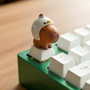 May include: A custom mechanical keyboard with white keycaps and a green frame. A capybara figurine wearing a chicken helmet sits atop a key. The keyboard is on a wooden surface.