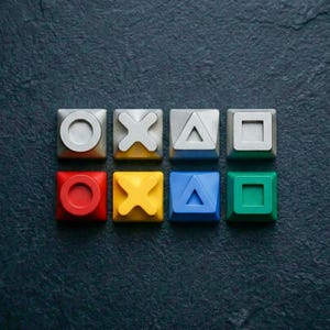 May include: Eight colorful square buttons with iconic gaming symbols: circle, X, triangle, square. The buttons are in red, yellow, blue, and green, with a dark background. A gaming-themed design.