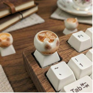 May include: A novelty keyboard keycap shaped like a miniature ceramic coffee cup with a latte art design. The keycap is white with brown detailing, set on a wooden keyboard base. Other similar keycaps are visible in the background.