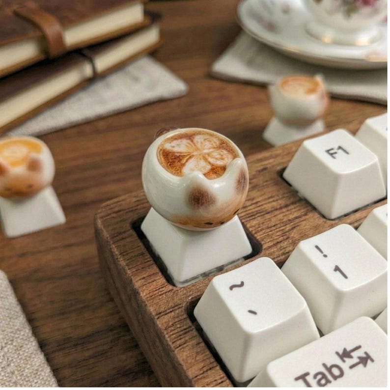 May include: A novelty keyboard keycap shaped like a miniature ceramic coffee cup with a latte art design. The keycap is white with brown detailing, set on a wooden keyboard base. Other similar keycaps are visible in the background.