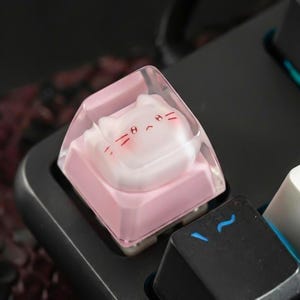 May include: A custom keyboard keycap featuring a pink base and a clear top with a cat face design. The cat face has red details for the eyes, nose, and whiskers. The keycap is set on a black keyboard.
