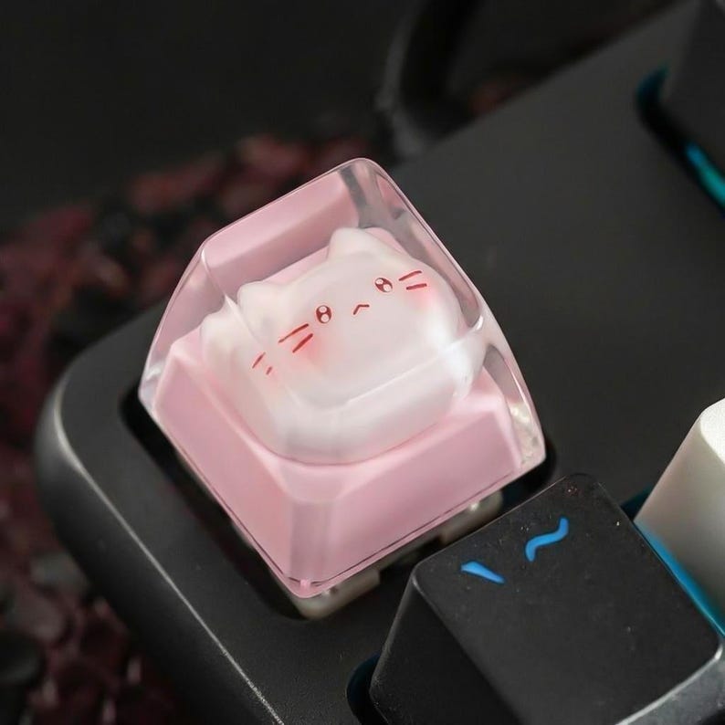 May include: A custom keyboard keycap featuring a pink base and a clear top with a cat face design. The cat face has red details for the eyes, nose, and whiskers. The keycap is set on a black keyboard.