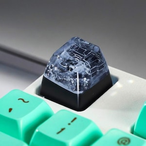 Può includere: Un copritasto personalizzato per tastiera con un design unico. Il copritasto ha una base nera e una parte superiore traslucida, che mostra un motivo marmorizzato bianco e grigio. Il copritasto è montato su una tastiera bianca.