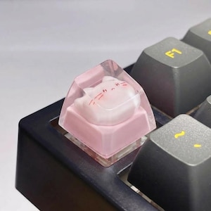 May include: A custom keyboard keycap featuring a pink cat design encased in clear resin. The keycap is pink and sits on a black keyboard. The cat has pink cheeks and whiskers. The keycap is a unique accessory for computer enthusiasts.