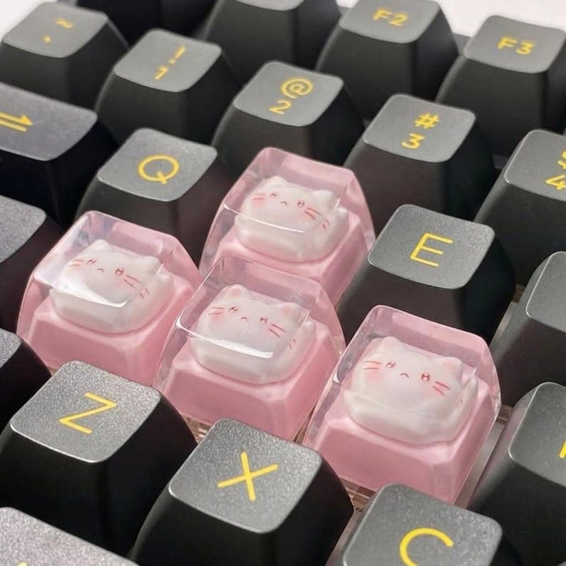 May include: Close-up of a mechanical keyboard with black keycaps and yellow lettering. Several keys are replaced with custom keycaps featuring a pink base and a clear top with a white cat face design.