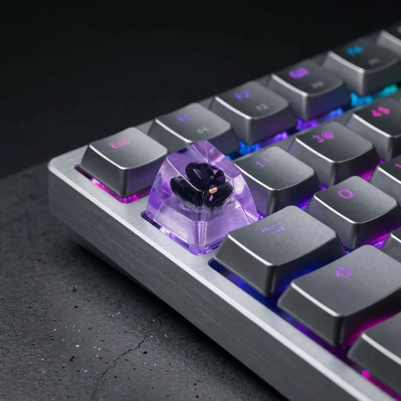 May include: Close-up of a mechanical keyboard with a custom keycap. The keycap is transparent purple with a dark floral design inside. The keyboard has gray keys and a silver frame, with purple and blue backlighting.