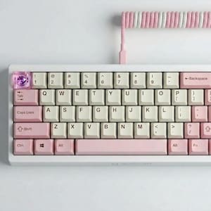 May include: A white mechanical keyboard with pink and cream-colored keycaps. The keyboard has a coiled cable with pink and white stripes. The keycaps are arranged in a standard QWERTY layout, with a few custom keycaps.