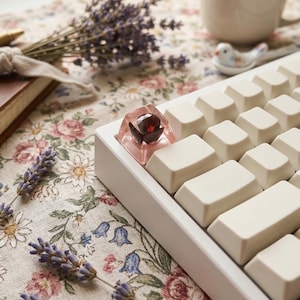 May include: A white mechanical keyboard with off-white keycaps. A custom keycap with a red gem is featured. The keyboard rests on a floral patterned fabric, with lavender sprigs and a book in the background.