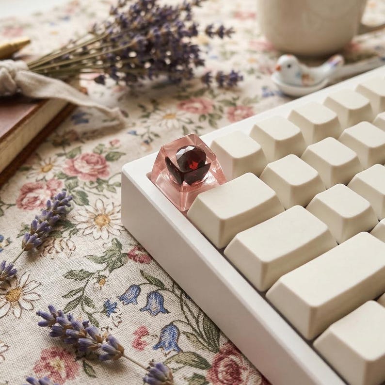 May include: A white mechanical keyboard with off-white keycaps. A custom keycap with a red gem is featured. The keyboard rests on a floral patterned fabric, with lavender sprigs and a book in the background.