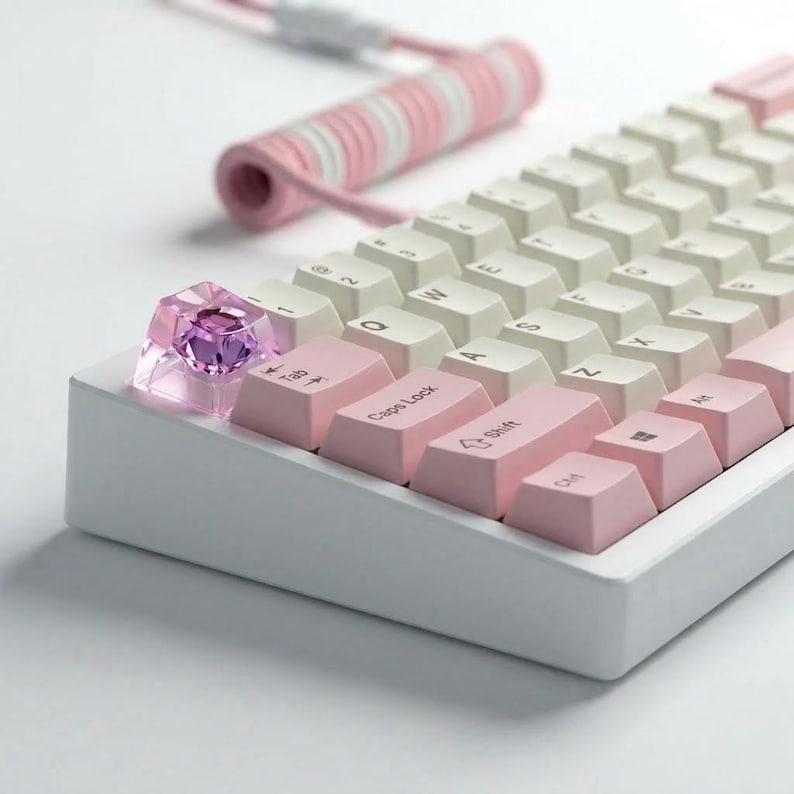 A white mechanical keyboard with pink and white keycaps. The keyboard has a custom keycap with a pink gem. A pink and white coiled cable is in the background.