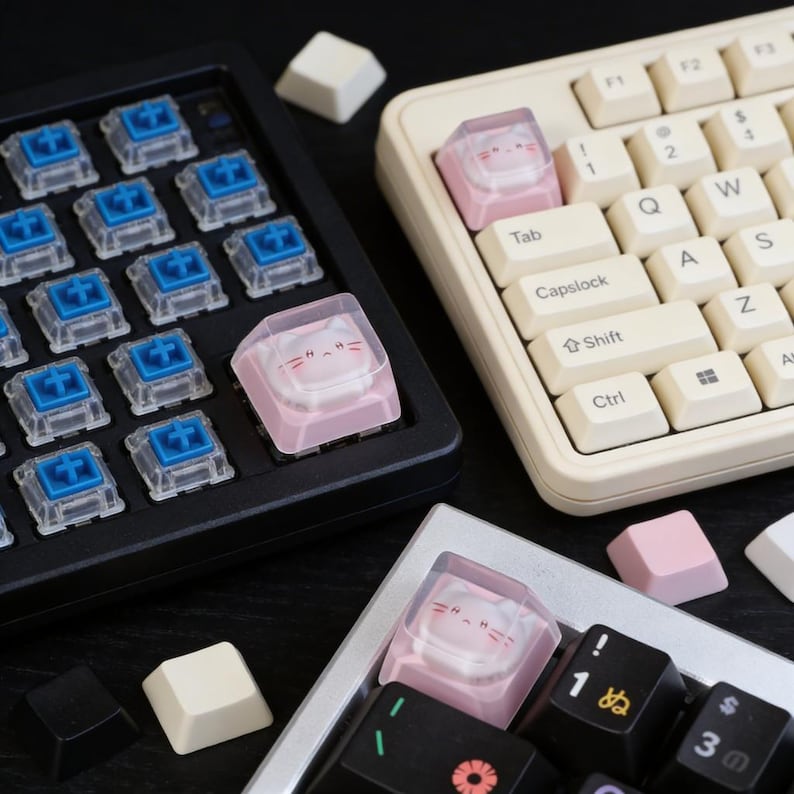 May include: Close-up of mechanical keyboard components. The image features a black keyboard base with blue switches, a cream-colored keyboard, and several keycaps. Some keycaps are pink with a cat face design. Other keycaps are white, black, and pink.