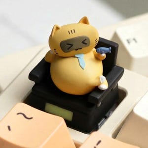Peut inclure: Figurine de chat jaune portant une cravate bleue et assis dans un fauteuil noir. Le chat tient un petit poisson bleu. La figurine est placée sur un clavier aux touches beiges.