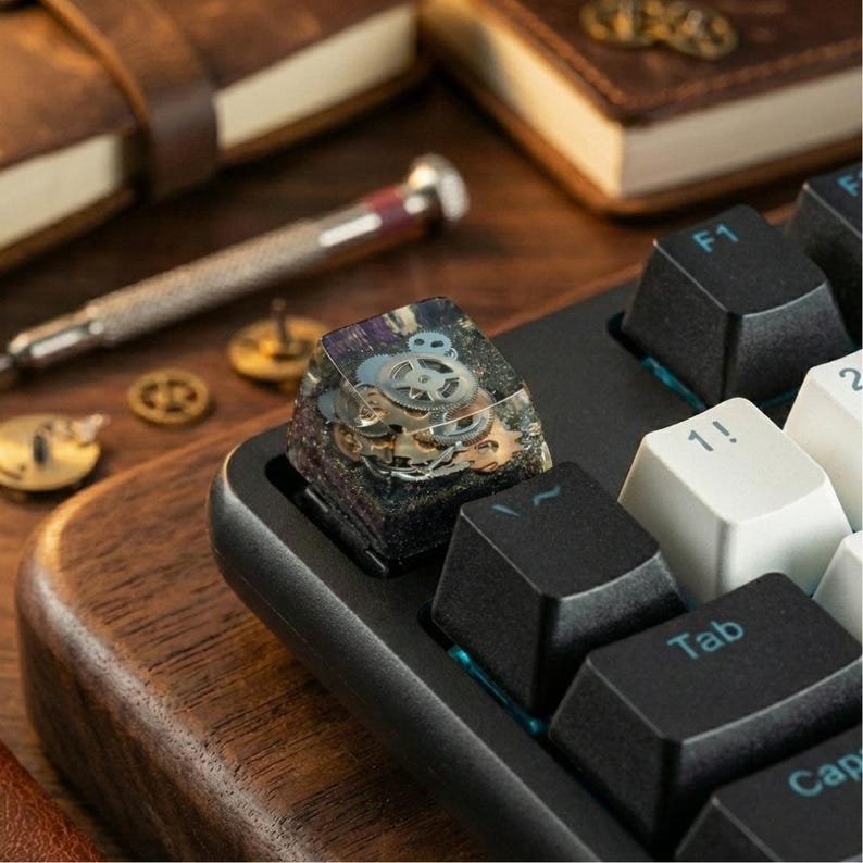 May include: Close-up of a mechanical keyboard with a custom keycap. The keycap is a clear resin block encasing watch gears and other small metal components. The keyboard has black keys and a wooden base. Other items on the wooden surface include a screwdriver and small metal pieces.