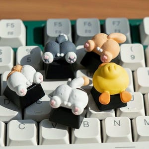 May include: Novelty keycaps in the shape of animal bottoms, in shades of grey, white, and yellow. The keycaps are on a white keyboard with green detailing. The keyboard keys are labelled.