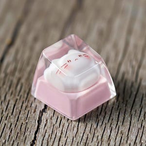 May include: A custom keyboard keycap featuring a white cat design with red accents encased in clear resin. The keycap sits on a pink base, creating a cute and unique aesthetic. The keycap is designed for mechanical keyboards.