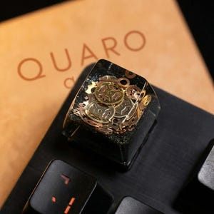 May include: A custom mechanical keyboard keycap with a steampunk design. The keycap is a clear resin block encasing brass gears and other mechanical components. The word "QUARO" is visible in the background.
