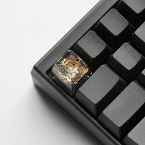 May include: Close-up of a black mechanical keyboard with a custom keycap. The keycap is clear with embedded gold-colored gears and mechanical components. The surrounding keys are black and rectangular.