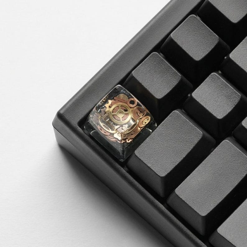 May include: Close-up of a black mechanical keyboard with a custom keycap. The keycap is clear with embedded gold-colored gears and mechanical components. The surrounding keys are black and rectangular.