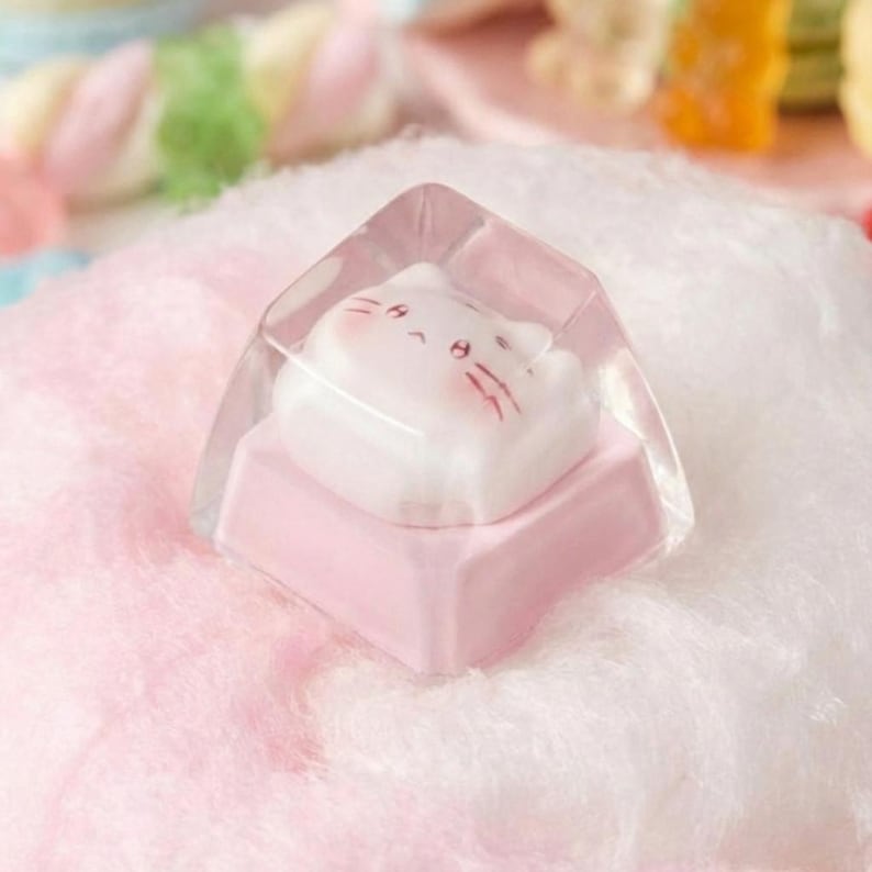 May include: A pink and clear cat-shaped keycap. The keycap features a white cat face with pink accents encased in a clear, geometric resin. The keycap sits on a pink base, resting on a fluffy pink surface.