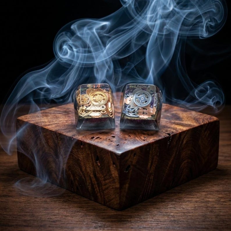 May include: Two rectangular, clear resin keycaps with intricate gear designs. One keycap features gold-colored gears, while the other has silver-colored gears. They rest on a dark wooden block, with wisps of smoke in the background.
