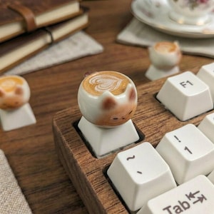 May include: A custom keyboard with cat-shaped keycaps. The keycaps are white with a brown and tan cat face design, and the keyboard has a wooden frame. The keycaps are shaped like a latte with a heart design.
