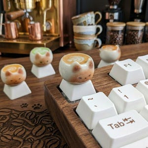 May include: A wooden keyboard with white keys and cat-shaped ceramic keycaps. The keycaps are designed to look like latte art. Other coffee-related items are in the background, including a coffee machine and cups.