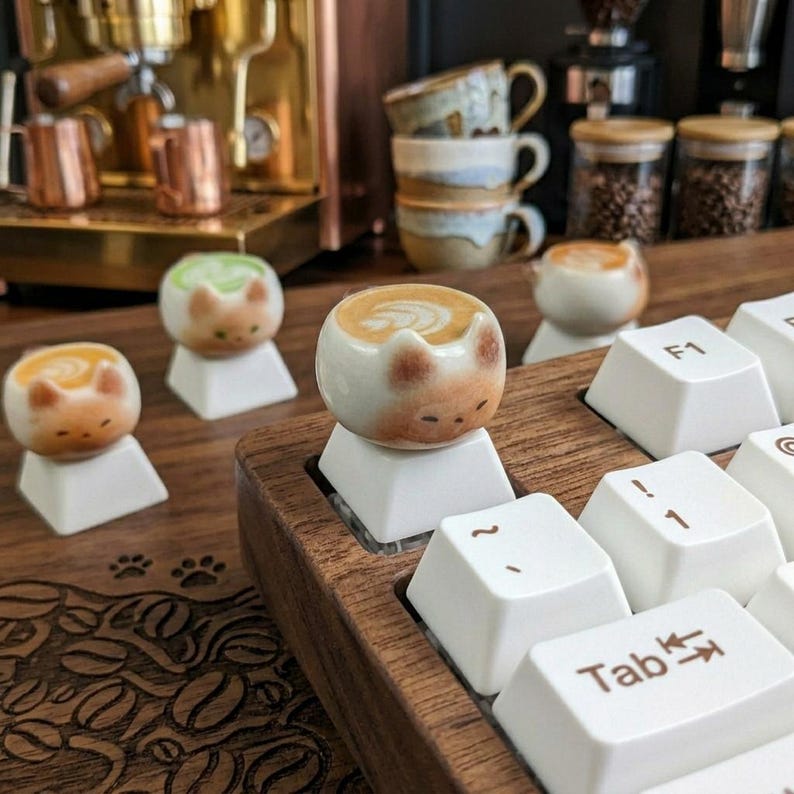 May include: A wooden keyboard with white keys and cat-shaped ceramic keycaps. The keycaps are designed to look like latte art. Other coffee-related items are in the background, including a coffee machine and cups.