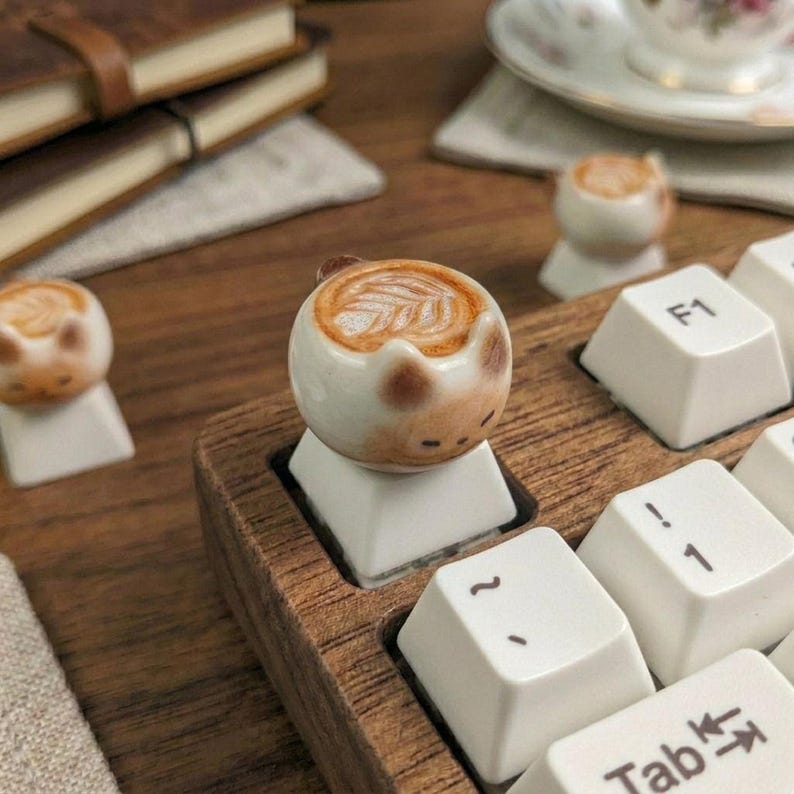 May include: A ceramic cat-shaped keycap with a latte art design, placed on a white keyboard key. The keycap is brown and white, featuring a coffee design. Other keycaps and a wooden keyboard frame are visible.