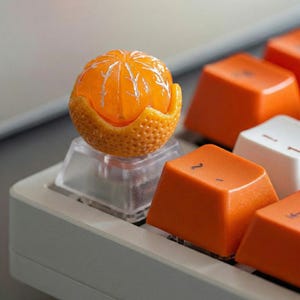 Puede incluir: Primer plano de un teclado mecánico con teclas naranjas y una tecla decorativa con forma de mandarina. La tecla naranja es una réplica detallada de una mandarina, con una cáscara texturizada y fruta segmentada.
