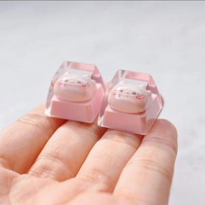 May include: Two custom keyboard keycaps. Each keycap features a pink base with a clear, geometric top. Inside each clear top is a light pink cat face with black whiskers and a small pink nose. The keycaps are held in a hand.