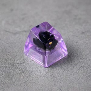 A custom keyboard keycap with a dark purple flower encased in transparent, light purple resin. The keycap has a geometric shape and is designed for mechanical keyboards. The flower has gold accents.