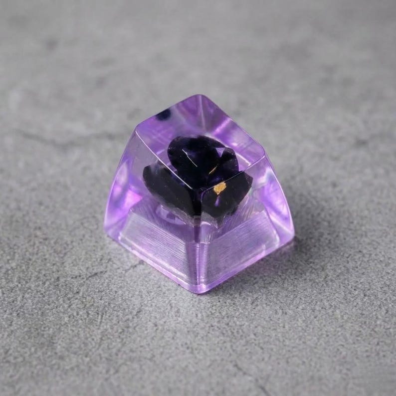 A custom keyboard keycap with a dark purple flower encased in transparent, light purple resin. The keycap has a geometric shape and is designed for mechanical keyboards. The flower has gold accents.
