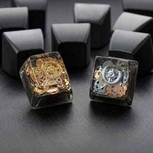 May include: Close-up of two artisan keycaps with embedded gears. One keycap features gold-colored gears, while the other has silver gears. Both are encased in a clear resin with a dark background. The keycaps are designed for mechanical keyboards.