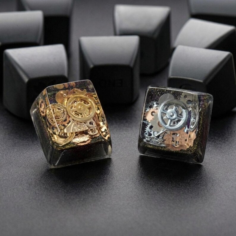 May include: Close-up of two artisan keycaps with embedded gears. One keycap features gold-colored gears, while the other has silver gears. Both are encased in a clear resin with a dark background. The keycaps are designed for mechanical keyboards.