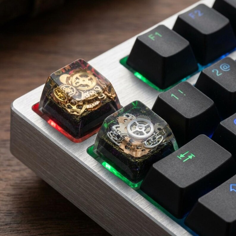 May include: Close-up of two custom mechanical keyboard keycaps. The keycaps are clear with embedded gold and silver gears. The keycaps are on a silver keyboard with black keys. One keycap has a red LED, the other has a green LED.