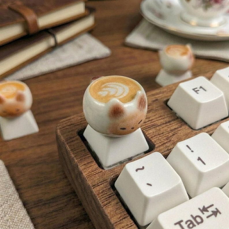 May include: A keyboard with custom cat-shaped keycaps, each featuring a latte art design. The keyboard has a wooden frame, and the keycaps are white. Other items in the background include notebooks and a teacup on a saucer.