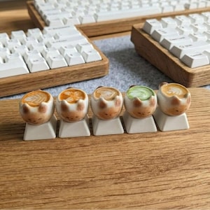 May include: Five ceramic cat-shaped keycaps, each with a different coffee art design, are displayed. The keycaps are on a wooden surface, with white keyboards in the background. The keycaps are a unique design.