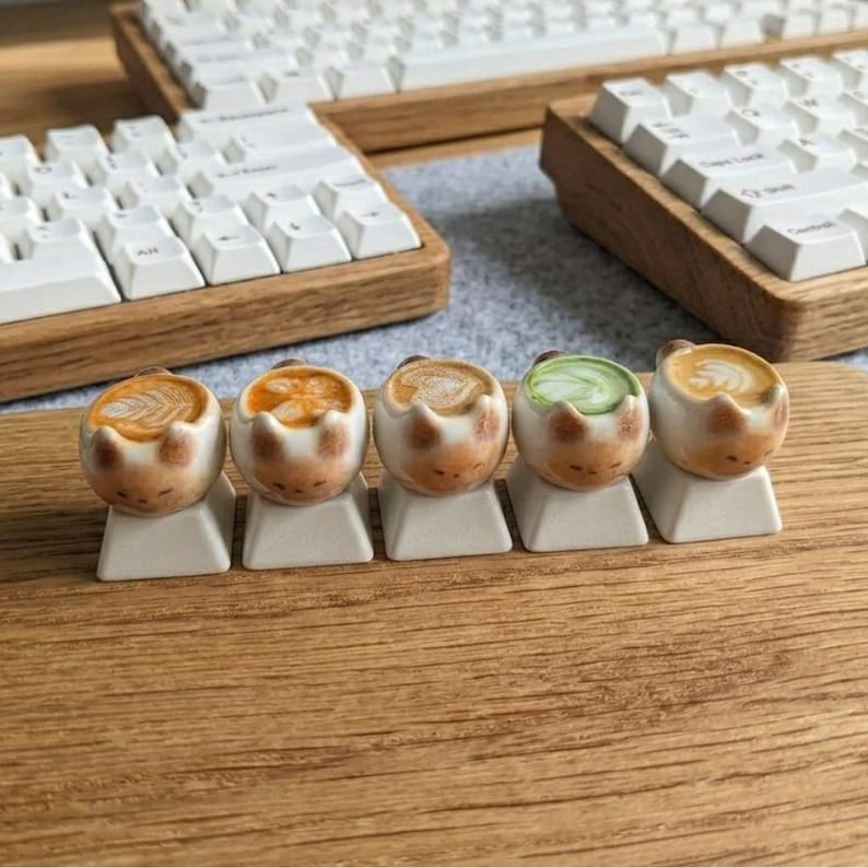 May include: Five ceramic cat-shaped keycaps, each with a different coffee art design, are displayed. The keycaps are on a wooden surface, with white keyboards in the background. The keycaps are a unique design.