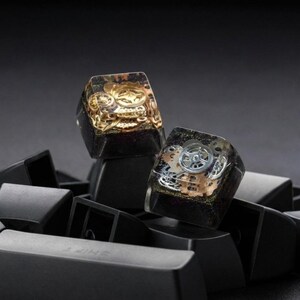 May include: Two custom mechanical keyboard keycaps featuring a clear resin top layer embedded with metallic gold and silver clockwork gears and cogs, set against a dark, glittery base, resting on a collection of plain black keycaps.