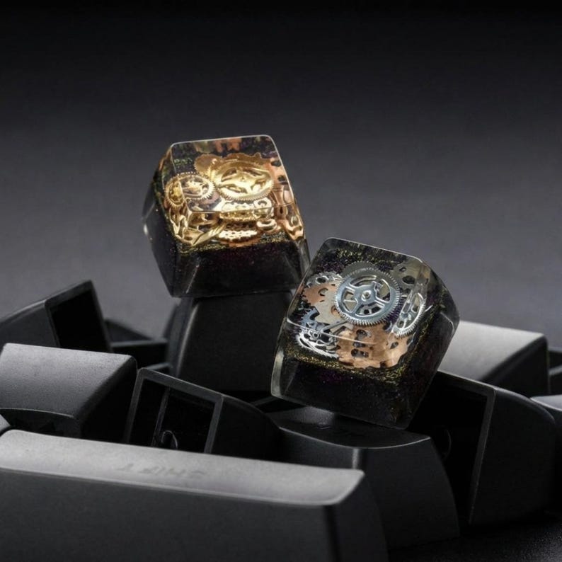 May include: Two custom mechanical keyboard keycaps featuring a clear resin top layer embedded with metallic gold and silver clockwork gears and cogs, set against a dark, glittery base, resting on a collection of plain black keycaps.