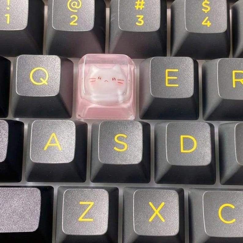 May include: Close-up of a mechanical keyboard with black keys and yellow lettering. A pink keycap with a cat face design is centered among the keys. The letters Q, E, A, S, D, Z, X, and C are visible.