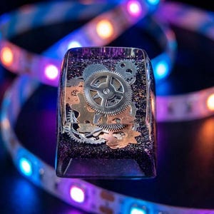 Könnte beinhalten: Ein rechteckiger, klarer Resin-Keycap mit eingebetteten silbernen und kupferfarbenen Zahnrädern. Der Keycap hat ein dunkles, glitzerndes Inneres und ist vor einem verschwommenen Hintergrund aus bunten LED-Lichtern platziert, was eine Steampunk-Ästhetik erzeugt.