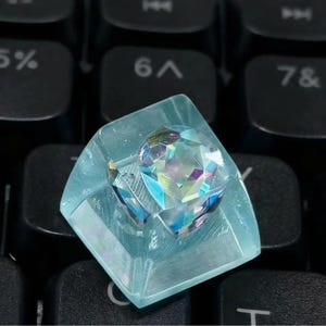 A custom keyboard keycap with a clear, light blue resin base. The keycap features a faceted, iridescent crystal centerpiece that reflects multiple colors. The keycap is set against a black keyboard.