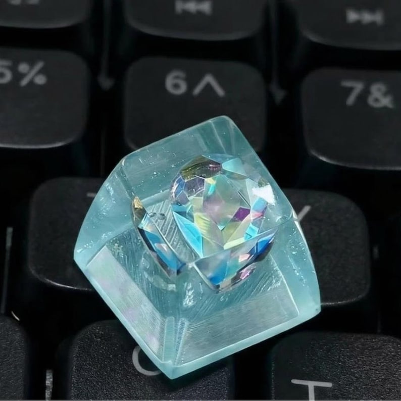 A custom keyboard keycap with a clear, light blue resin base. The keycap features a faceted, iridescent crystal centerpiece that reflects multiple colors. The keycap is set against a black keyboard.