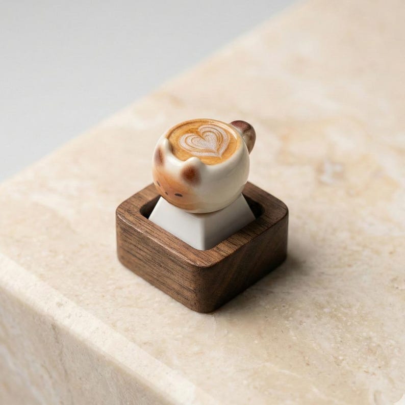 May include: A novelty ceramic coffee cup featuring latte art in a heart shape, set on a white square within a dark wooden base. The cup has brown details and a small handle. The base is square and made of dark wood.