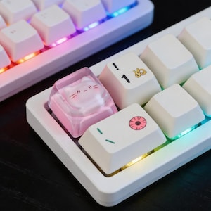 May include: Close-up of a white mechanical keyboard with custom keycaps. One keycap is a clear pink cat design. Other keycaps have symbols and illustrations. The keyboard has a colorful LED backlight.