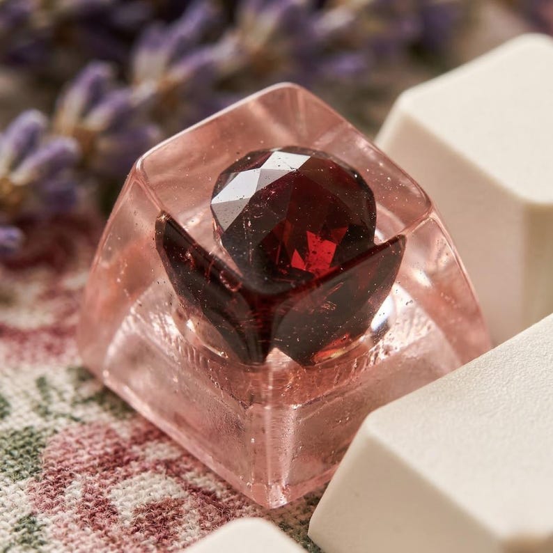 A custom keycap with a faceted, deep red gemstone center, encased in a clear, pink resin. The keycap has a flower-like design and is surrounded by white keyboard keys. The background includes floral fabric and lavender sprigs.