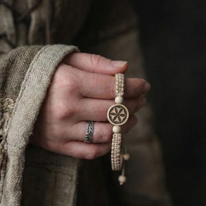 Può includere: Un braccialetto in macramè beige con un ciondolo circolare in legno con un disegno a stella intagliata. Il braccialetto è tenuto in una mano, con un anello d'argento al dito. Lo sfondo è in una tonalità neutra e attenuata.