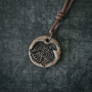 May include: A circular, brown pendant featuring a carved bird motif, hanging from a brown leather cord. The pendant has a textured finish and a metal ring for the cord. The backdrop is a dark, textured surface.