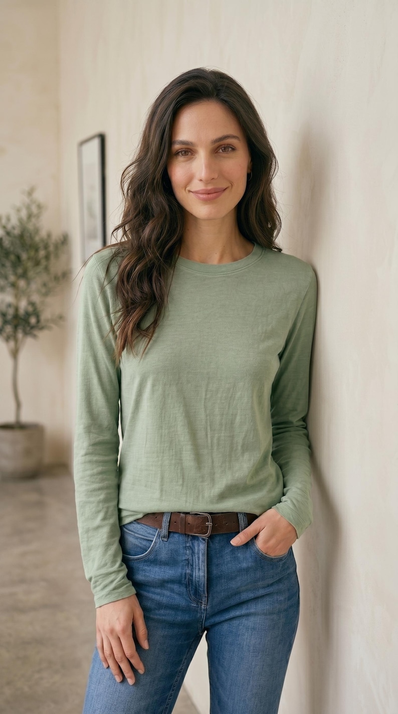 May include: A long-sleeved, light green shirt paired with blue jeans and a brown belt. The model is leaning against a neutral-colored wall. The shirt has a crew neck and a relaxed fit, suitable for casual wear.